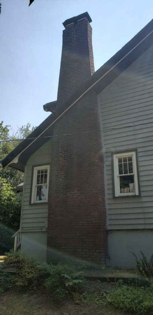 A tall Portland chimney masonry on the side of a grey house with white-trimmed windows.