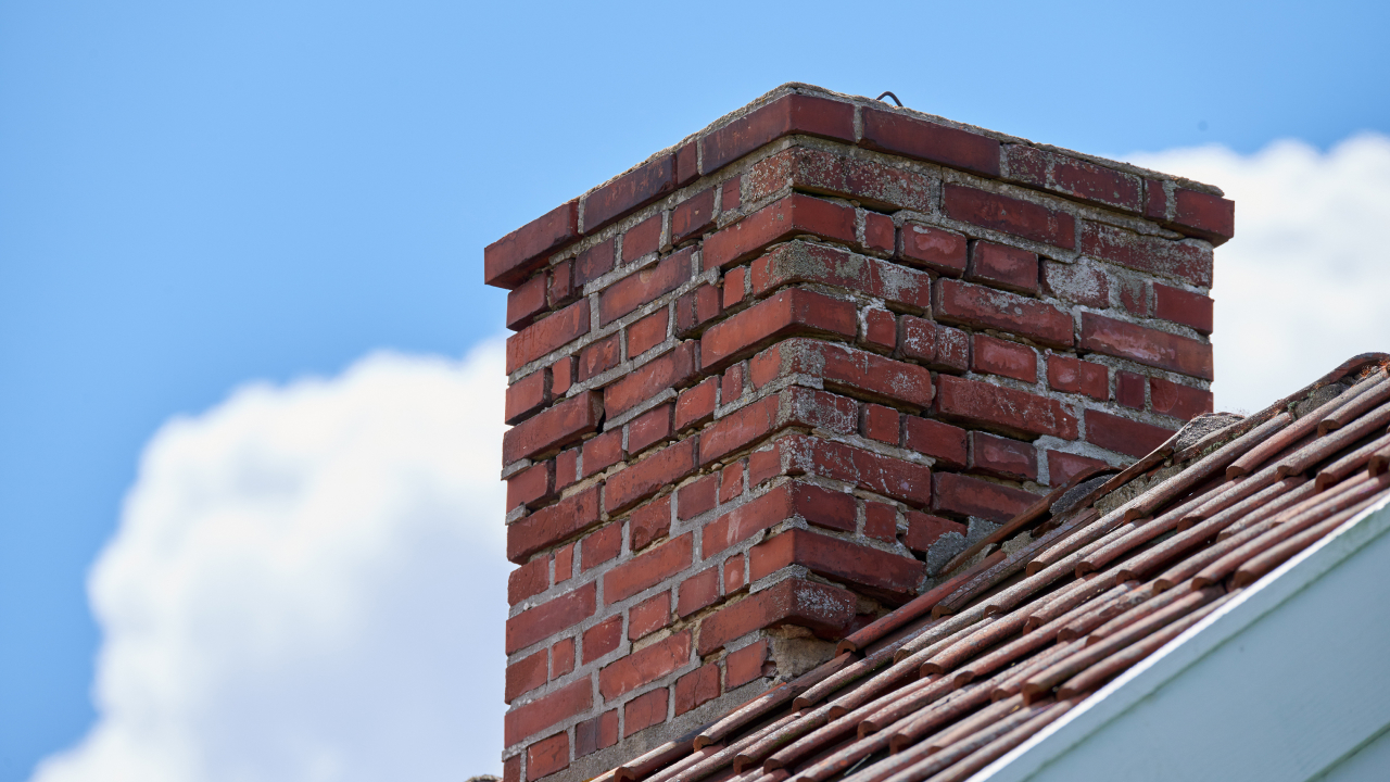 Close-up of a brick chimney on top of a house roof with a blue sky and scattered clouds in the background.