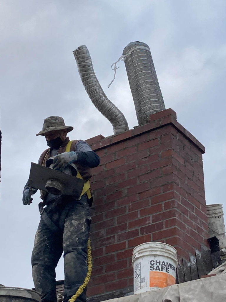 A construction worker in protective gear is working on a brick chimney; he is holding a tool, and there are metal ducts and a white bucket labeled "Chamber Safe" nearby.