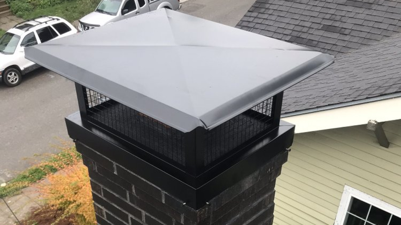 A rectangular chimney cap with a metal top and mesh sides is installed on a brick chimney, with cars and a house roof visible in the background.
