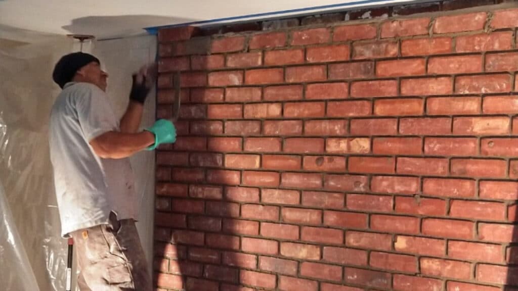 Worker installing or repairing a brick wall indoors, wearing gloves and a head covering.