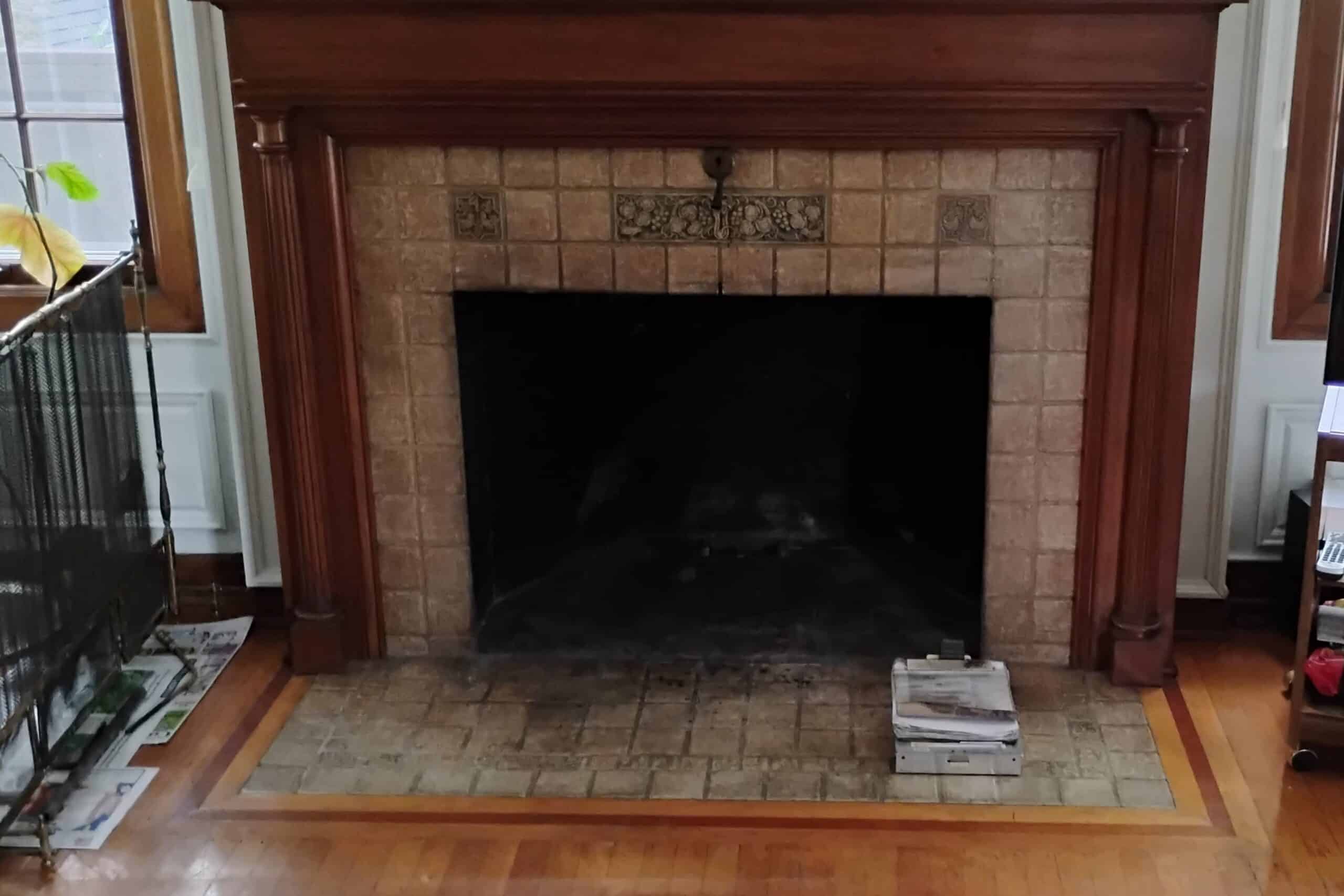 A tiled fireplace with a wooden mantle, surrounded by wooden flooring. A stack of newspapers is visible on the tiled hearth.