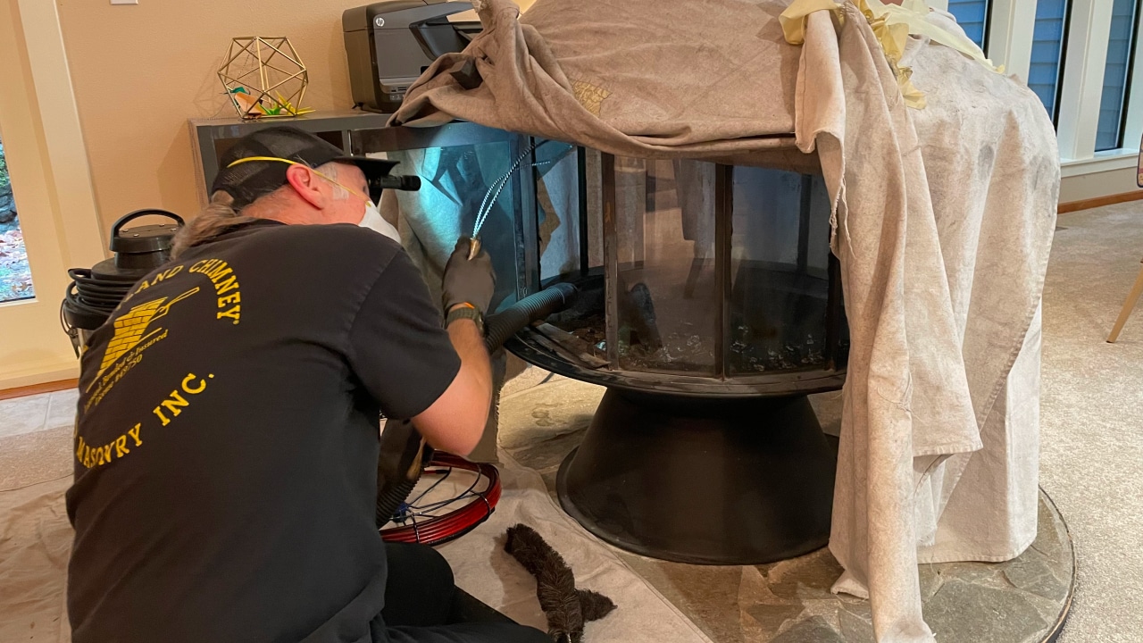 Technician cleaning and inspecting a fireplace chimney inside a home, wearing gloves and safety glasses, with tools and cloths nearby.