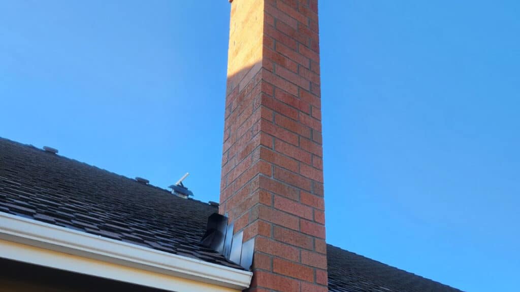 Red brick chimney extending from a dark shingled roof against a clear blue sky.