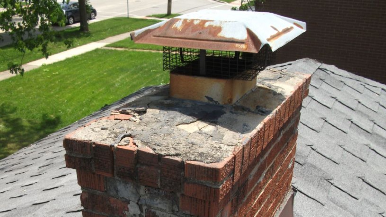 A deteriorating brick chimney with a rusted metal cap and cracked mortar is shown, surrounded by grey roof shingles. A street and grass are visible in the background.