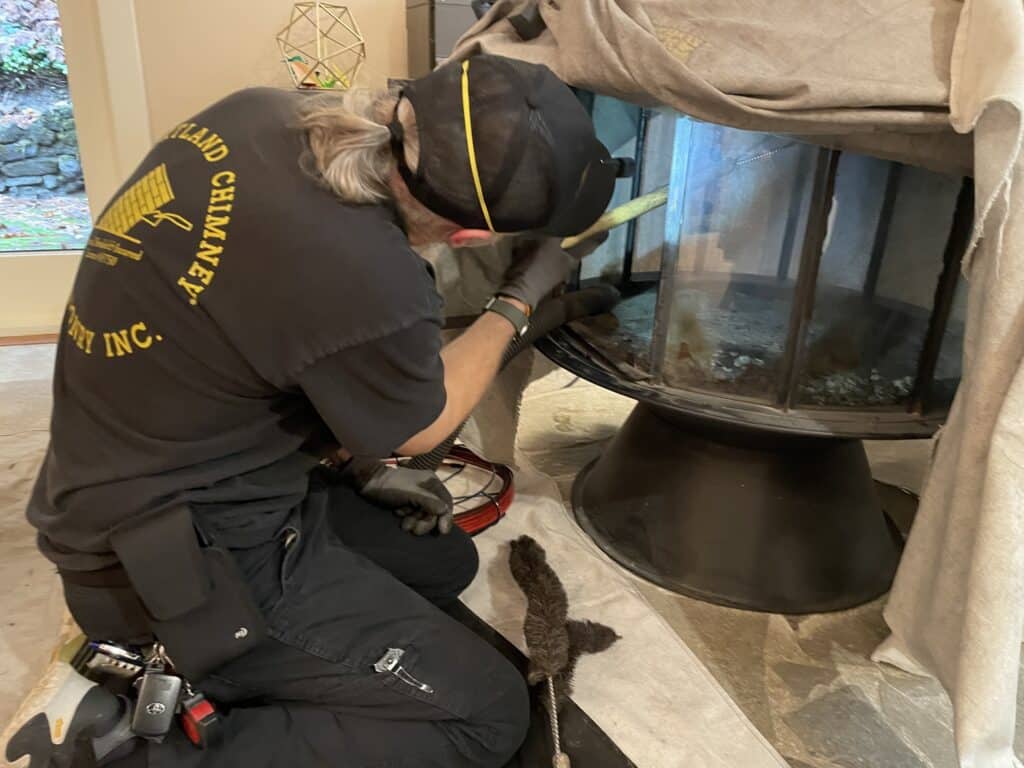 A person wearing a black shirt and cap cleans a large glass fireplace, surrounded by tools and a partially covered area.