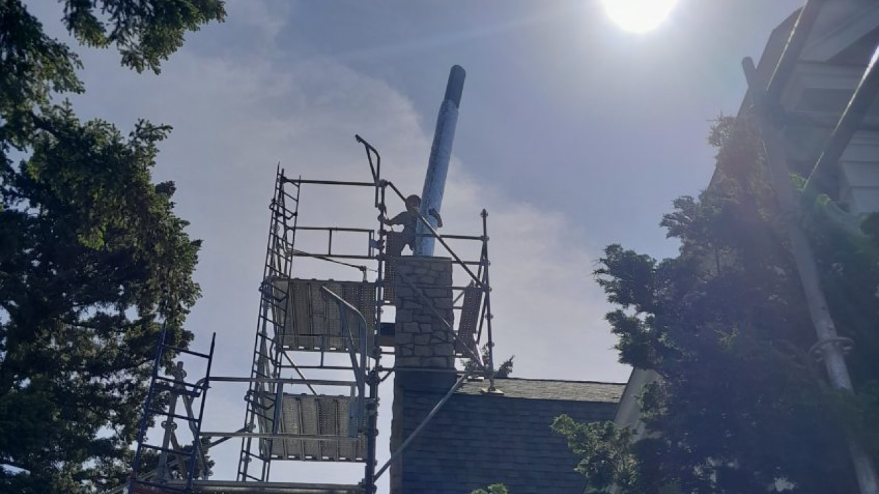 Scaffolding surrounds a chimney under repair on a sunny day, with trees visible nearby.