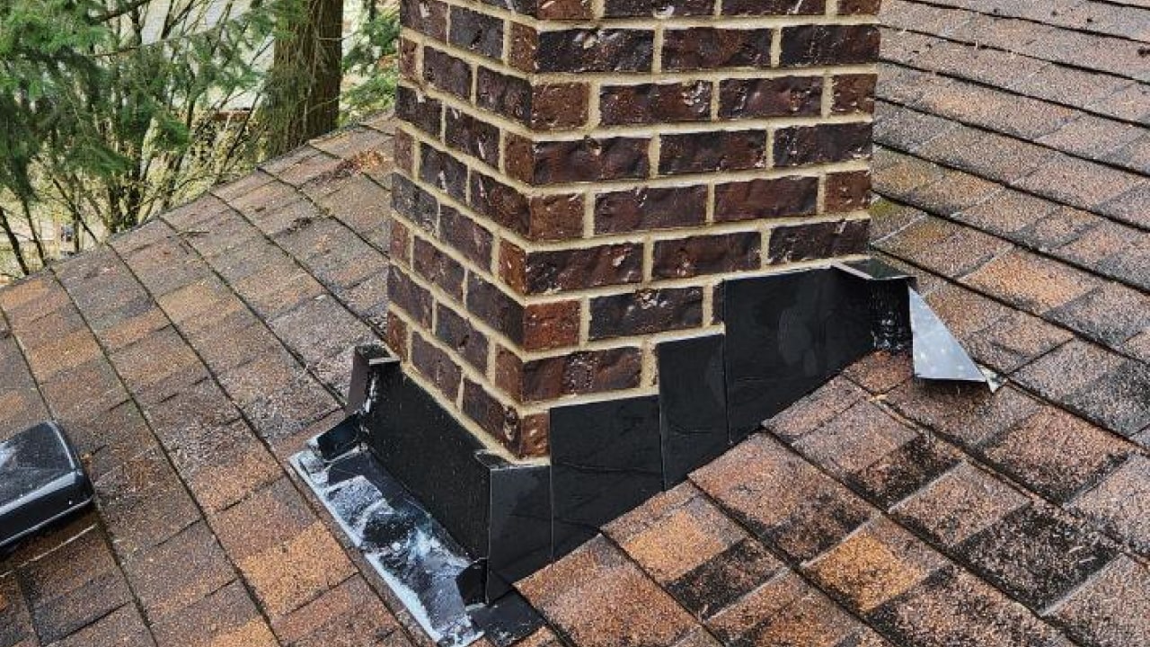 Brick chimney with black flashing on a brown shingle roof, surrounded by trees in the background.
