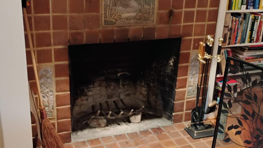A brick fireplace with a blackened interior. Decorative tiles on the sides. Nearby are a broom, brass fireplace tools, and a bookcase filled with books.