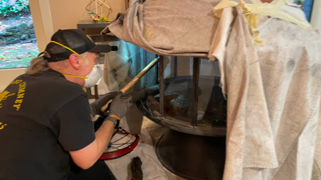 Person cleaning a fireplace with a brush, wearing a cap, mask, and gloves. The fireplace is covered with sheets, with cleaning tools nearby.
