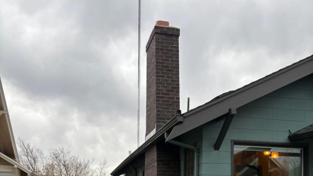 A grey house with a tall brick chimney against a cloudy sky. A window reflects light below the roof.