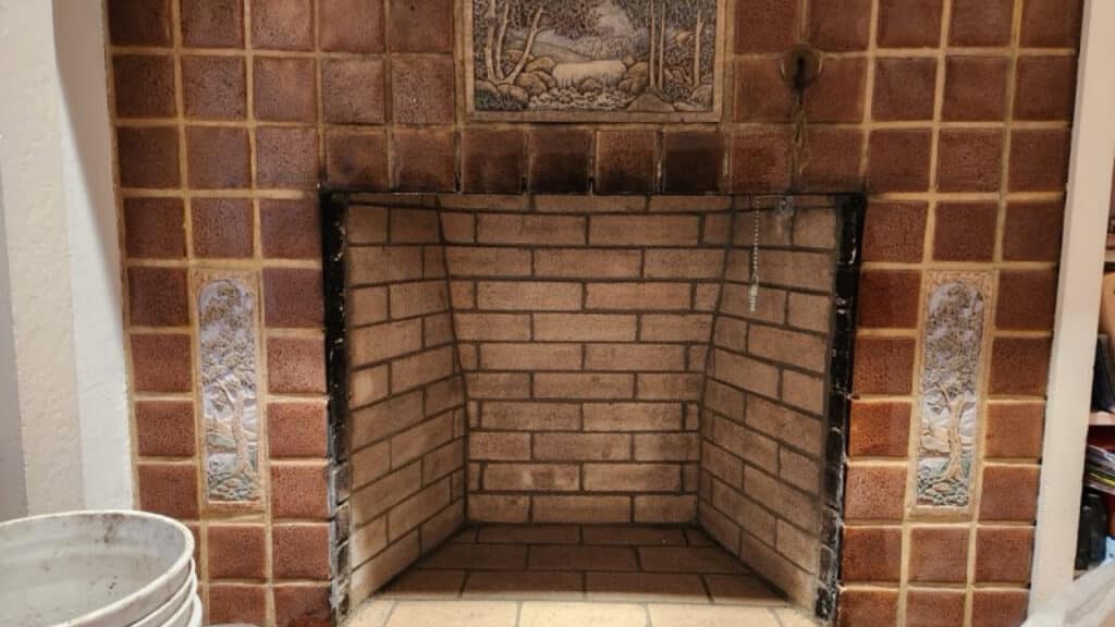 A brick fireplace with decorative tiled surround, featuring nature-themed reliefs, and an empty white plastic bucket to the left.