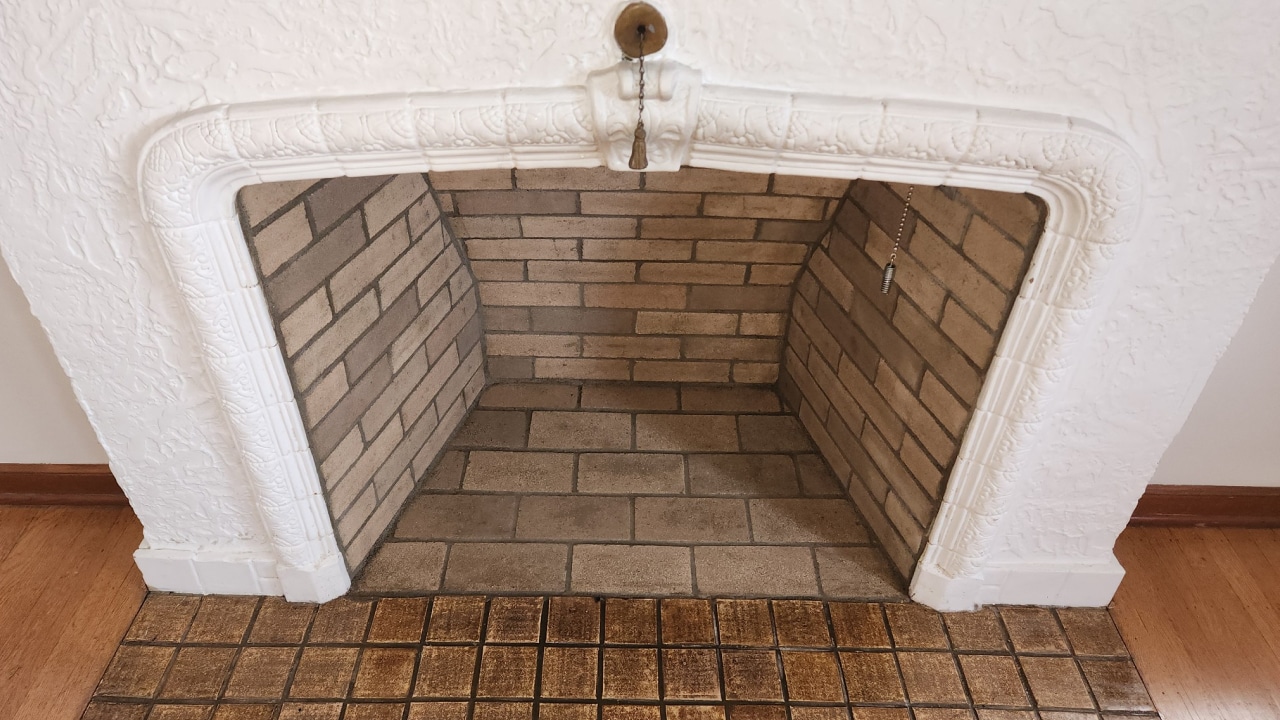 An empty fireplace with white decorative molding, brick interior, and a small chain hanging from the top. The hearth is tiled with square, brown tiles.