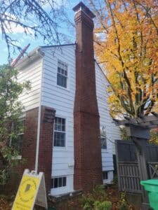 Two-story white house with a tall brick chimney, trees nearby displaying autumn colors, and a 