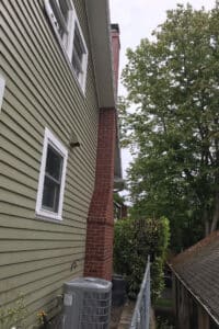 Side view of a house with green siding and white-framed windows, featuring a red brick chimney. An air conditioning unit sits beside a chain-link fence, with trees visible in the background.