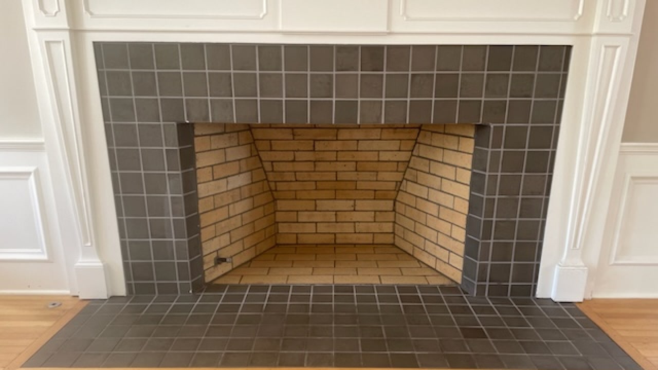 Fireplace with a black tiled exterior and a brick-lined interior. Light wooden floor and white wall panels surround the fireplace.