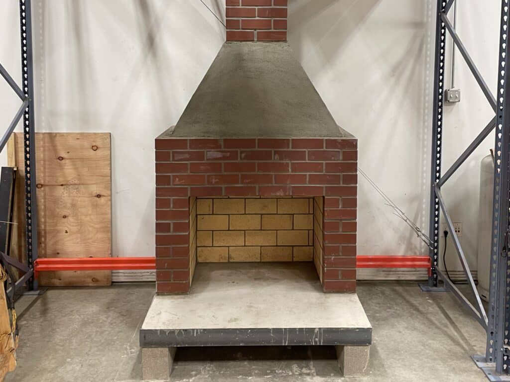 A brick and concrete fireplace structure stands between metal shelves in an industrial setting.