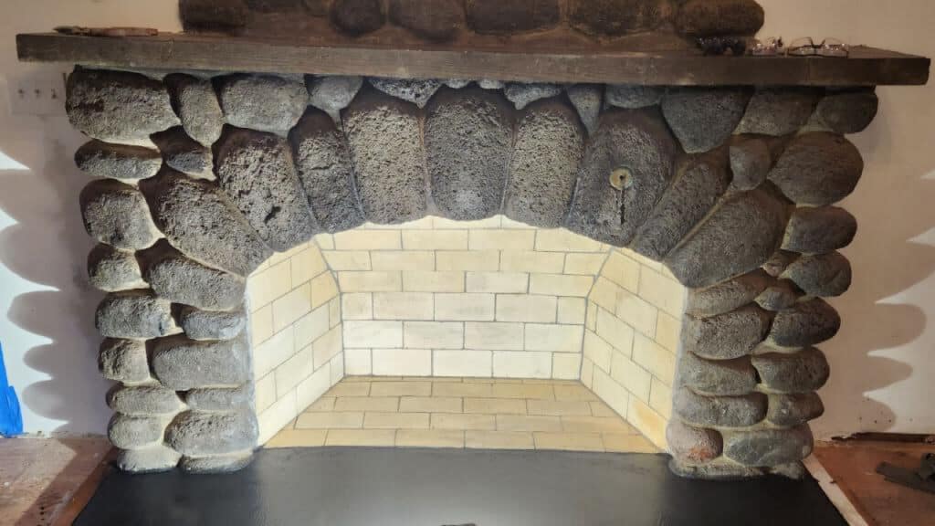 Stone fireplace with a rounded arch, surrounded by large, rough rocks. The interior features light brickwork, and the hearth is dark and flat.