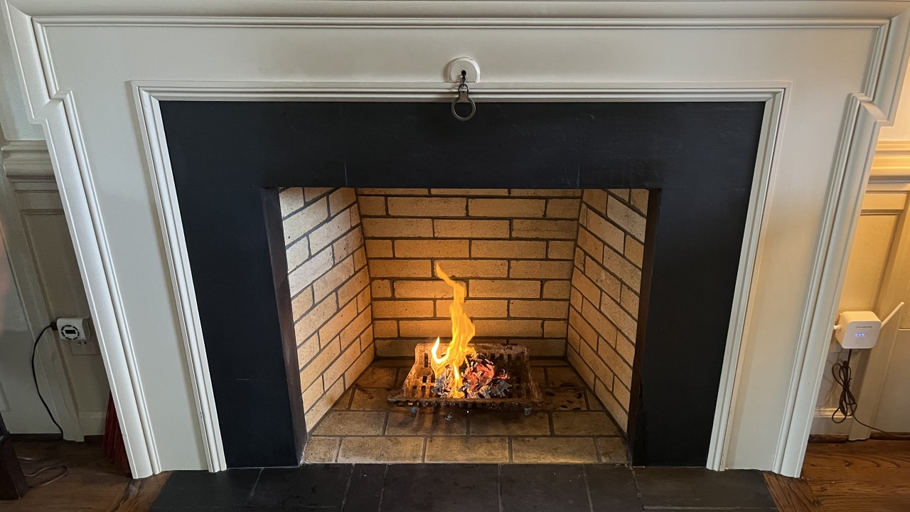 A lit fireplace with a small flame burning on logs inside a black-framed, white mantled hearth surrounded by light brick.