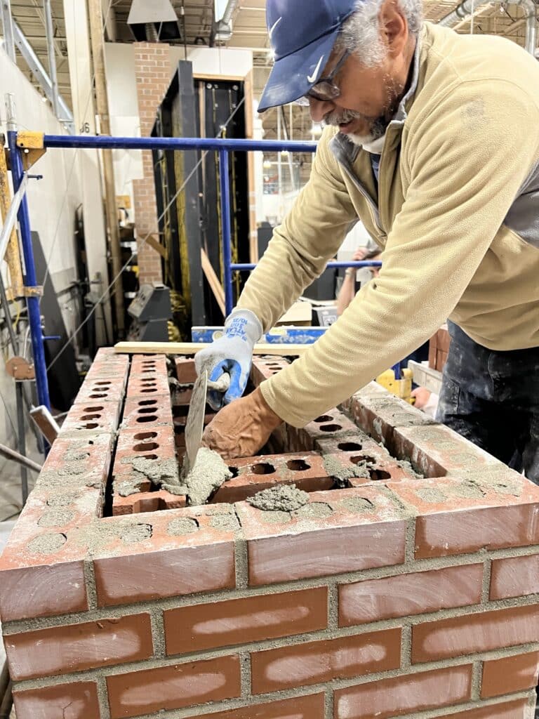 A man wearing gloves and a cap is applying mortar to red bricks while building a brick wall indoors.