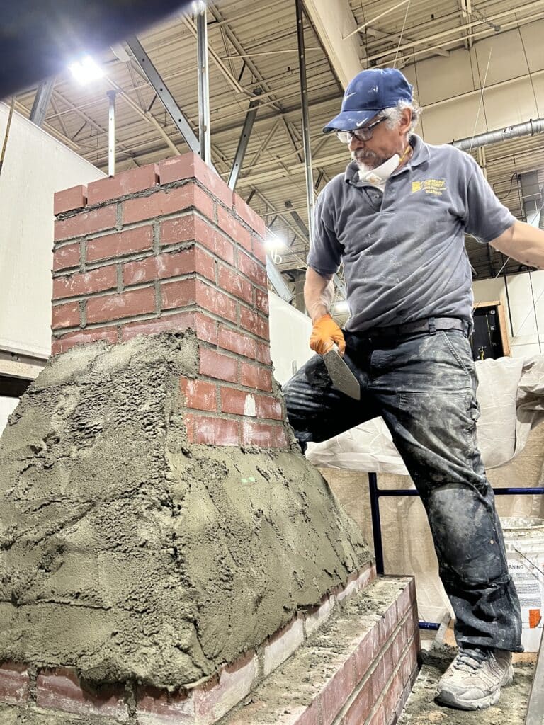 A man wearing work clothes and a cap applies mortar to a brick structure inside a building using a trowel.