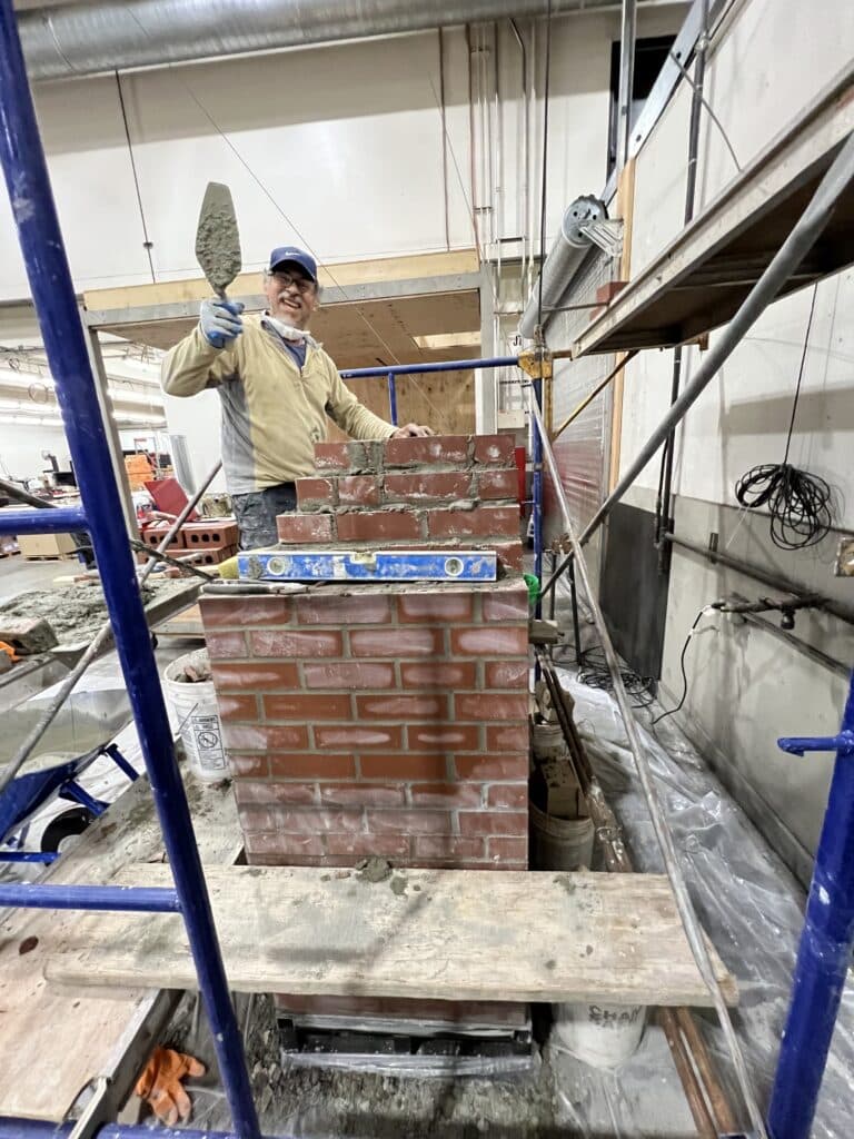A person in work clothes and gloves is constructing a brick wall indoors, holding a trowel and smiling at the camera. Construction tools and materials are visible around.