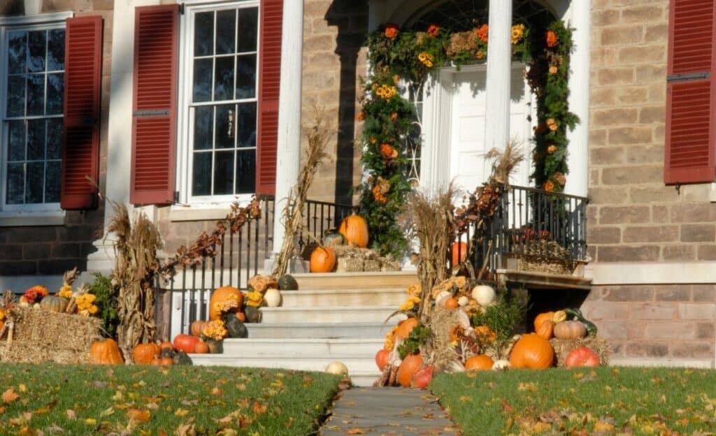 A house entrance decorated for autumn with pumpkins, gourds, cornstalks, and a garland of leaves and flowers around the door.