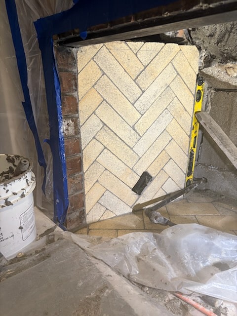 A section of a wall with light-colored tiles arranged in a herringbone pattern, construction tools and plastic sheets are visible around the work area.