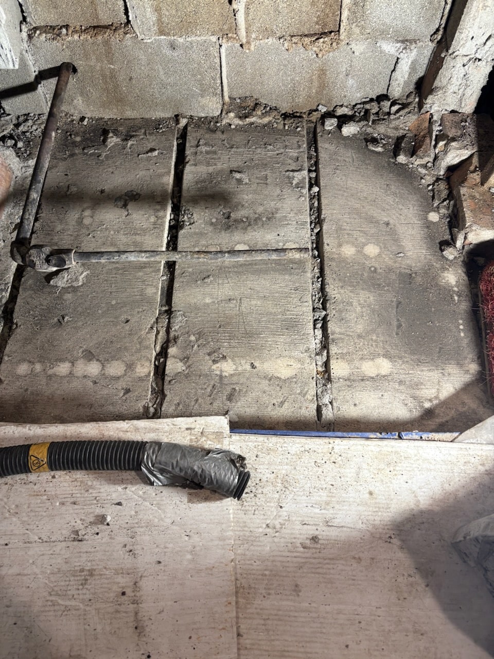 A close-up of a concrete floor section with metal bars and exposed cinder blocks in the background; a black corrugated tube lies in the foreground.