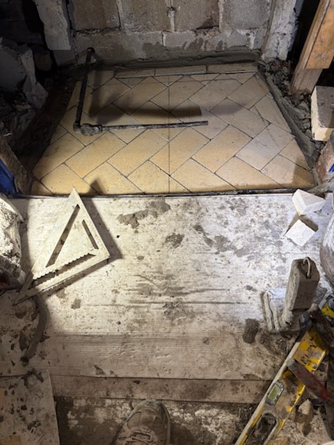 A partially tiled floor with beige tiles laid in a herringbone pattern, surrounded by construction tools and debris in a work area.
