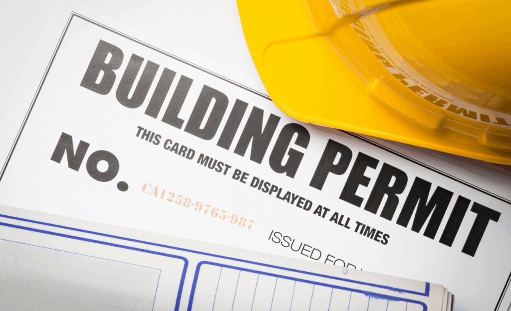 A building permit document and a yellow hard hat placed on a desk; the permit displays a permit number and instructions for display.