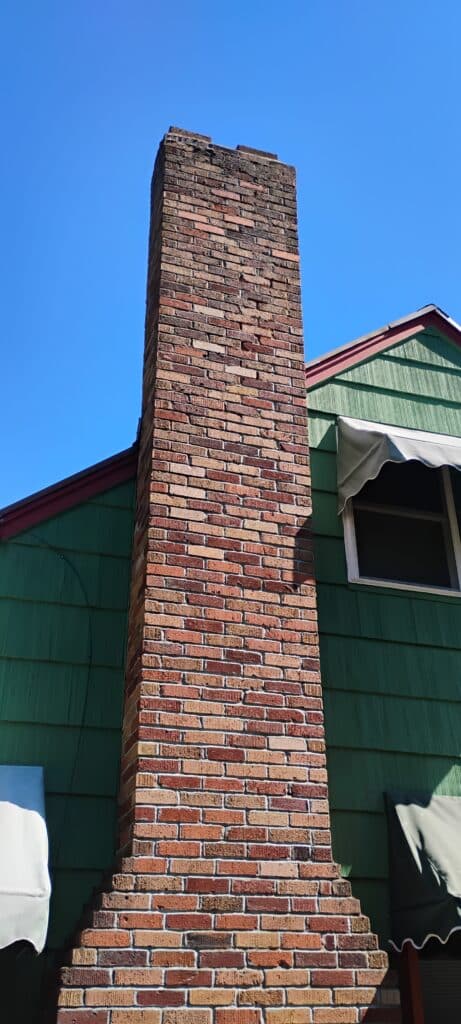A tall, rectangular brick chimney attached to a green house with white window and door awnings, under a clear blue sky.