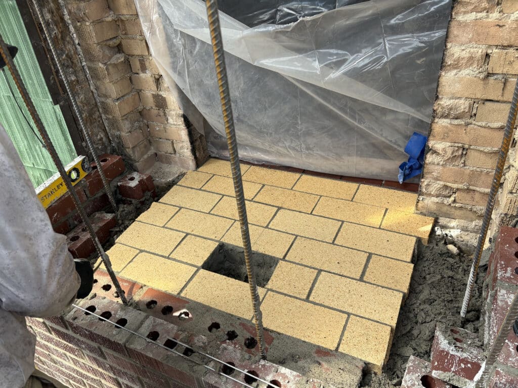 A partially constructed brick wall with yellow bricks and a square hole in the center, surrounded by concrete and building materials.