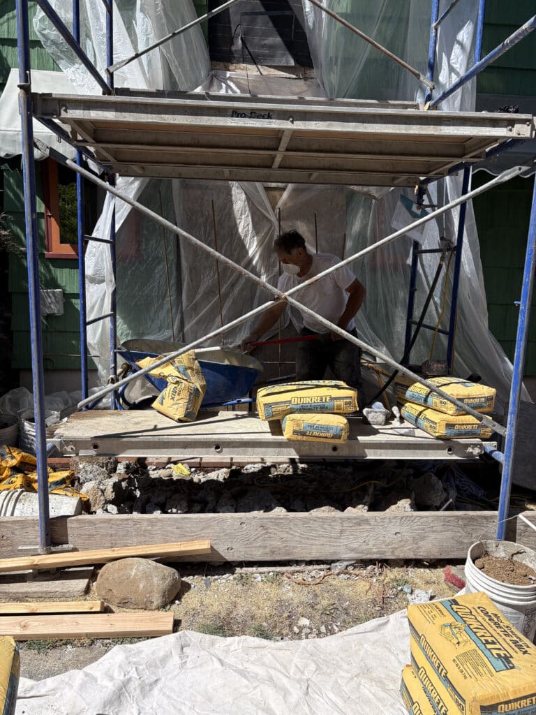 A construction worker mixes cement under scaffolding, surrounded by bags of Quikrete and rubble, at a building site covered with plastic sheeting.