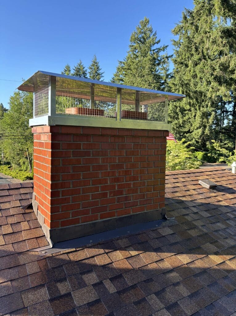 A red brick chimney with a metal mesh cap on a shingled roof, surrounded by tall green trees under a clear blue sky.