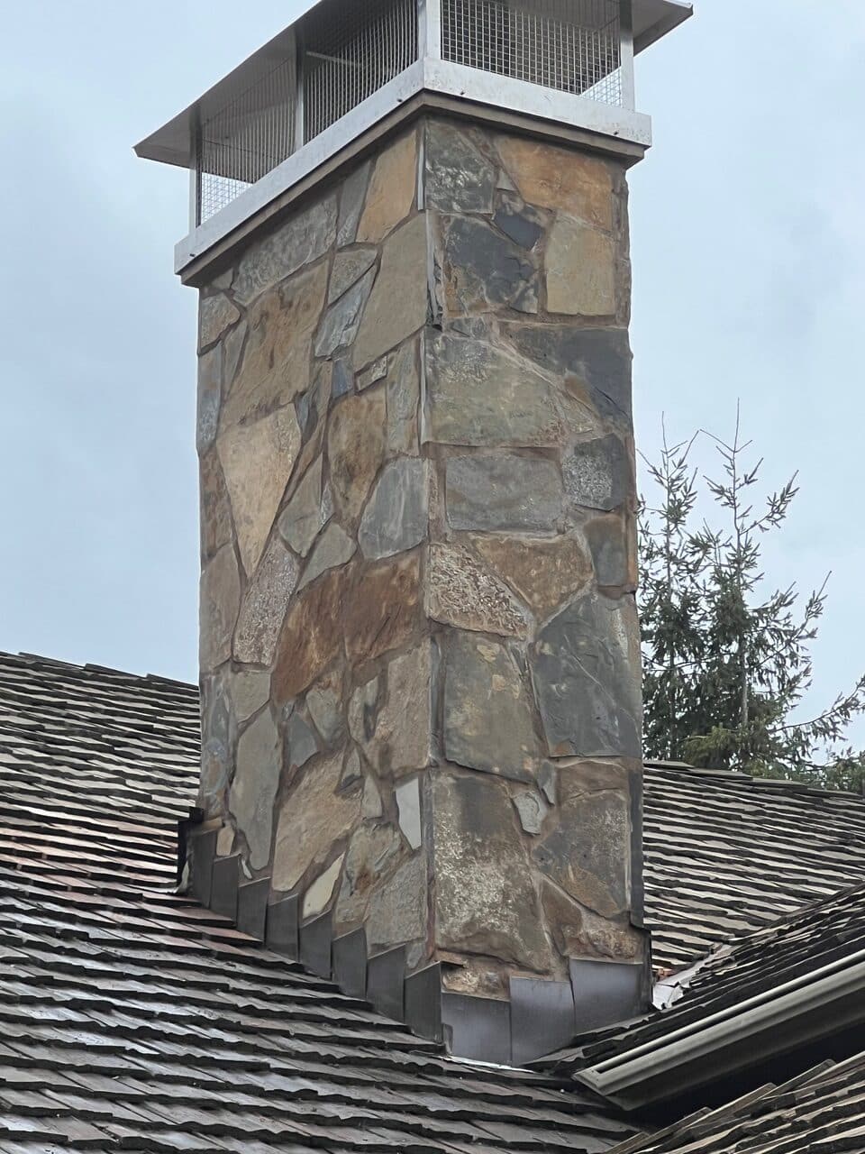 A stone chimney with a metal cap extends above a shingled roof on a cloudy day.