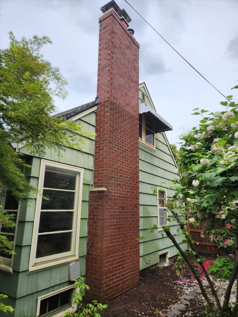 A tall, red brick chimney is attached to the exterior of a green, two-story house, with trees and bushes surrounding the yard.