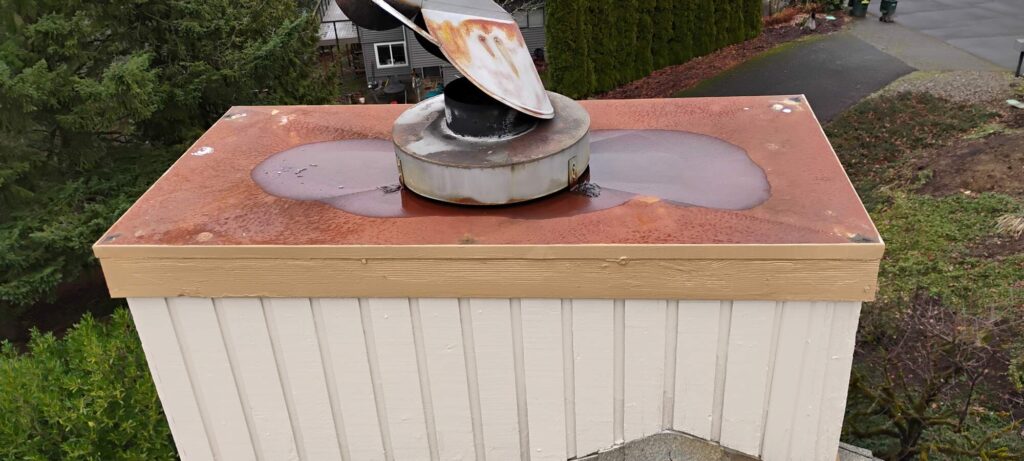 A rusted metal chimney cap with large water stains sits atop a white wooden structure next to a road and greenery.