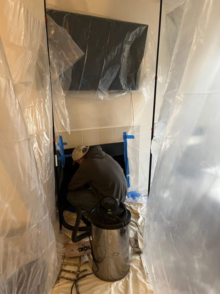A person in a hoodie and cap works on a fireplace surrounded by plastic sheeting, protective covers, and a shop vacuum.