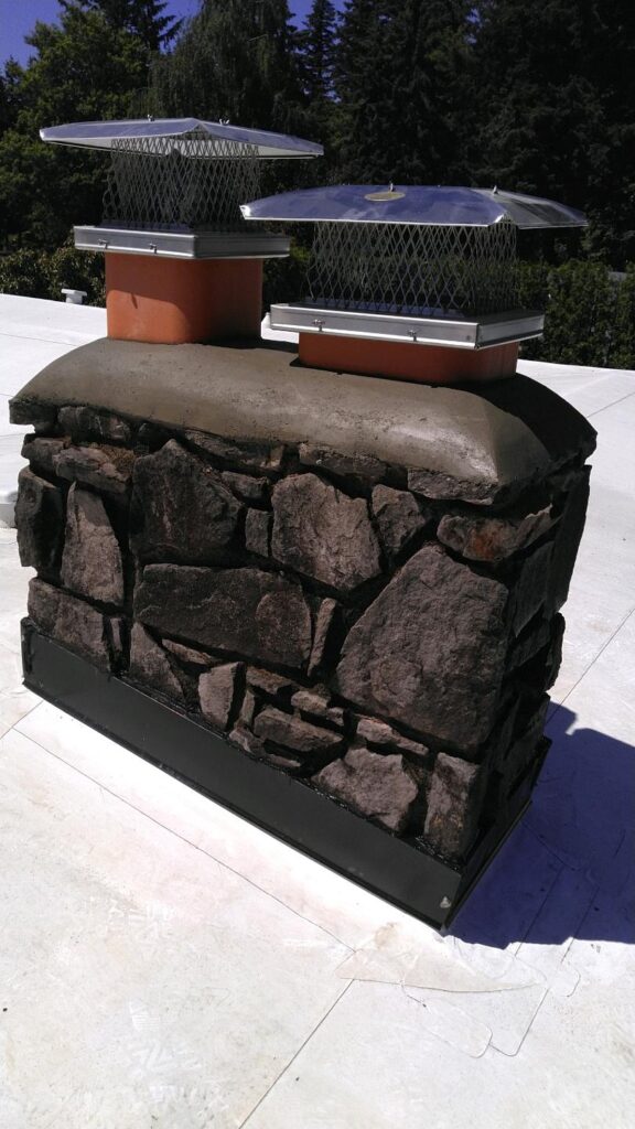 Stone chimney with two metal chimney caps on a flat white roof; trees and greenery are visible in the background.