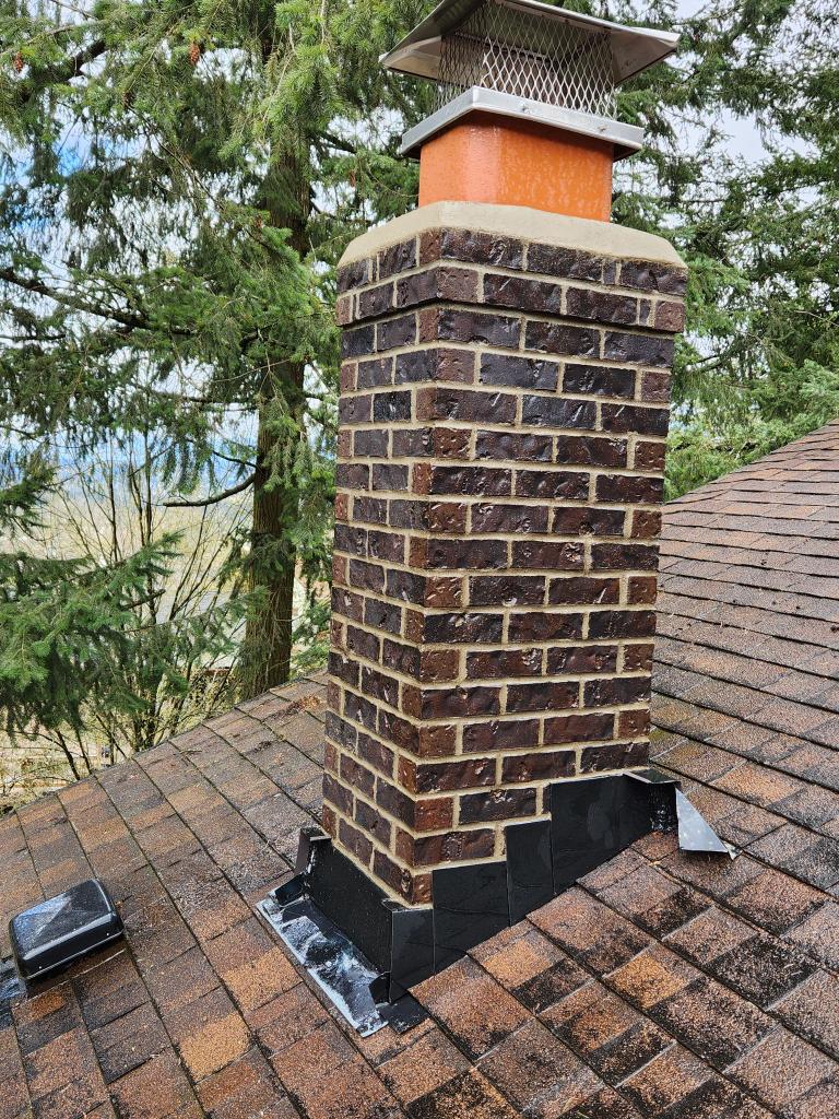 A brick chimney with a metal cap rises from a shingled roof, surrounded by black flashing and trees in the background.