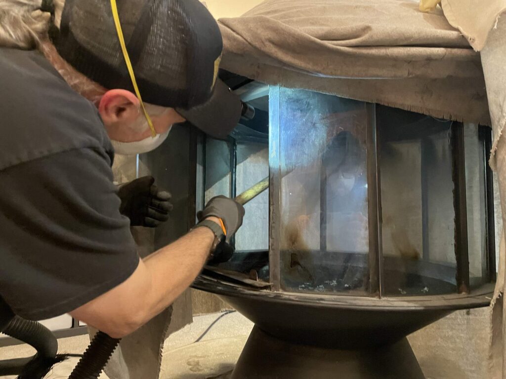 Person wearing gloves and a mask cleans the inside of a metal fireplace insert with a tool, with a cloth draped over the top.