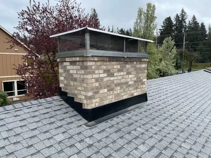 A brick chimney with a metal mesh cap sits on a shingled roof in Portland, surrounded by trees and a nearby building, under an overcast sky.