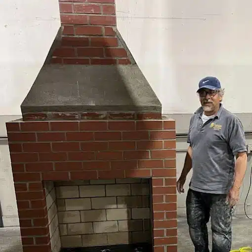 A man wearing work clothes stands next to a large, unfinished indoor brick fireplace structure inside a workshop or industrial space.