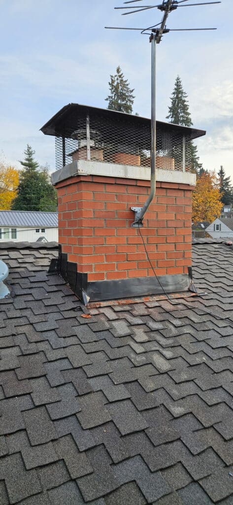 A brick chimney with a metal cap and attached TV antenna rises from an asphalt shingle roof, surrounded by houses and trees in the background.